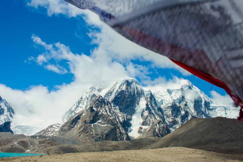 High wind blowing prayer flags beside Gurudongmar Lake with snow-covered mountains in backdrop